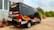 A black minibus with red and yellow graphics is parked outside a building with large windows. The vehicle has contact information and logos, and there are decorative plants in red pots nearby. In the background, there are trees and a clear sky.