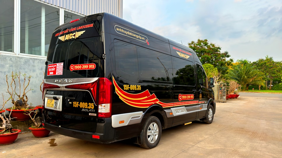 A black minibus with red and yellow graphics is parked outside a building with large windows. The vehicle has contact information and logos, and there are decorative plants in red pots nearby. In the background, there are trees and a clear sky.