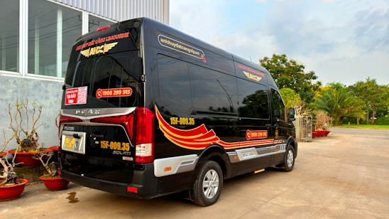 A black minibus with red and yellow graphics is parked outside a building with large windows. The vehicle has contact information and logos, and there are decorative plants in red pots nearby. In the background, there are trees and a clear sky.