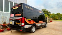 A black minibus with red and yellow graphics is parked outside a building with large windows. The vehicle has contact information and logos, and there are decorative plants in red pots nearby. In the background, there are trees and a clear sky.