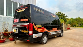 A black minibus with red and yellow graphics is parked outside a building with large windows. The vehicle has contact information and logos, and there are decorative plants in red pots nearby. In the background, there are trees and a clear sky.