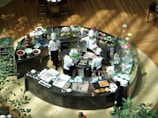 A group of chefs wearing white uniforms and hats are working in a circular kitchen station, preparing various foods. The area is filled with a variety of ingredients and dishes, including fruits, chocolate fountains, and pastries. The surrounding area has a wooden floor and a few plants.