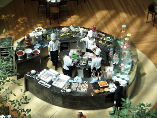 A group of chefs wearing white uniforms and hats are working in a circular kitchen station, preparing various foods. The area is filled with a variety of ingredients and dishes, including fruits, chocolate fountains, and pastries. The surrounding area has a wooden floor and a few plants.