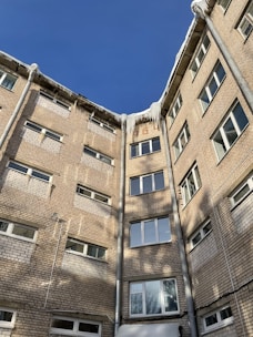 A brick residential building with multiple stories is shown with icicles hanging from the rooftop. The sun casts shadows on the walls, and the sky is clear and blue. Several windows are visible, and drainpipes run down the building.