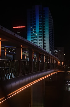 A nighttime cityscape featuring a high-rise building illuminated with blue lighting. In the foreground, an elevated walkway with orange lights creates a leading line toward the building. Bright red signage with Chinese characters adds a hint of vibrancy amid the darkness.