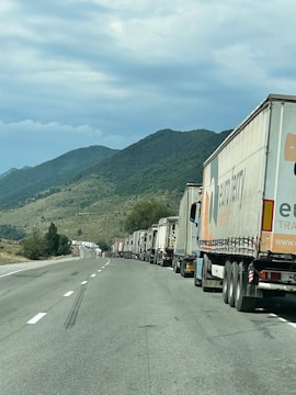 A long line of trucks is parked on a highway. The road stretches into the distance, bordered by rolling hills covered with green vegetation. The sky is cloudy, suggesting overcast weather. One truck is prominently visible with the branding 'euroberry' on its side.