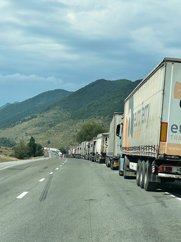 A long line of trucks is parked on a highway. The road stretches into the distance, bordered by rolling hills covered with green vegetation. The sky is cloudy, suggesting overcast weather. One truck is prominently visible with the branding 'euroberry' on its side.