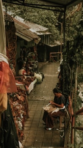 A woman sitting on a small stool is weaving or making handicrafts in an outdoor market setting. The area is surrounded by lush greenery and a variety of traditional crafts and artifacts displayed on both sides of the walkway. The scene suggests a rustic atmosphere with earthy tones.