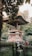 A traditional Balinese structure with an ornate, carved roof situated amidst lush green foliage. The building features intricate woodwork and a stone base, showcasing cultural architectural details. A wooden ladder leans against the structure, and the surrounding area is rich with tropical plants and trees.
