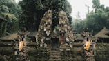 An ancient stone gate, adorned with moss and surrounded by dense tropical vegetation. Traditional Balinese architectural elements are visible, including ornate carvings and cloth decorations. The scene is set in a misty and lush forest environment, creating a serene and mystical atmosphere.