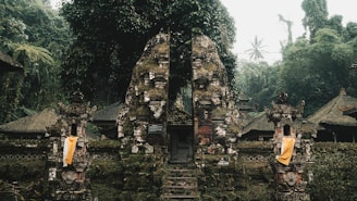 An ancient stone gate, adorned with moss and surrounded by dense tropical vegetation. Traditional Balinese architectural elements are visible, including ornate carvings and cloth decorations. The scene is set in a misty and lush forest environment, creating a serene and mystical atmosphere.