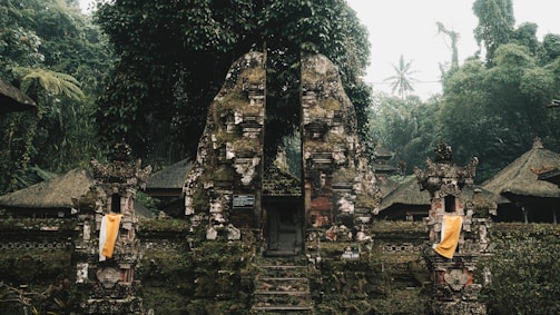 An ancient stone gate, adorned with moss and surrounded by dense tropical vegetation. Traditional Balinese architectural elements are visible, including ornate carvings and cloth decorations. The scene is set in a misty and lush forest environment, creating a serene and mystical atmosphere.