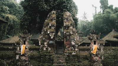 An ancient stone gate, adorned with moss and surrounded by dense tropical vegetation. Traditional Balinese architectural elements are visible, including ornate carvings and cloth decorations. The scene is set in a misty and lush forest environment, creating a serene and mystical atmosphere.