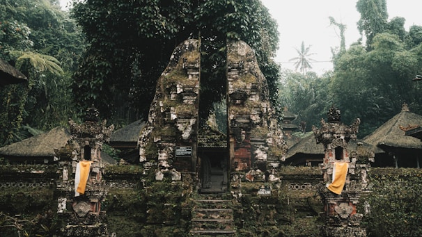 An ancient stone gate, adorned with moss and surrounded by dense tropical vegetation. Traditional Balinese architectural elements are visible, including ornate carvings and cloth decorations. The scene is set in a misty and lush forest environment, creating a serene and mystical atmosphere.