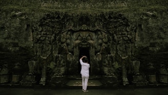 A person stands before an ancient, intricately carved stone entrance, likely part of a temple or historical structure. The carvings depict various figures and ornate patterns, with a dark opening leading inside.