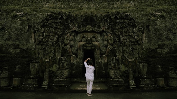 A person stands before an ancient, intricately carved stone entrance, likely part of a temple or historical structure. The carvings depict various figures and ornate patterns, with a dark opening leading inside.