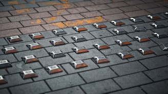 A paved surface made of interlocking bricks with rectangular reflectors embedded in a grid pattern. The bricks vary in color, ranging from dark gray to light brown, and there is some yellow paint marking on the surface.