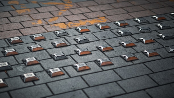A paved surface made of interlocking bricks with rectangular reflectors embedded in a grid pattern. The bricks vary in color, ranging from dark gray to light brown, and there is some yellow paint marking on the surface.