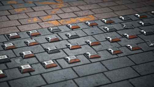 A paved surface made of interlocking bricks with rectangular reflectors embedded in a grid pattern. The bricks vary in color, ranging from dark gray to light brown, and there is some yellow paint marking on the surface.