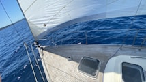 The image captures the deck of a sailboat cutting through deep blue waters, with the sun casting shadows on the wooden surface. The sail is fully extended, adding an element of motion and adventure. In the background, a faint line of the shoreline can be seen along with a clear blue sky.