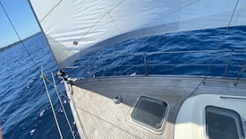 The image captures the deck of a sailboat cutting through deep blue waters, with the sun casting shadows on the wooden surface. The sail is fully extended, adding an element of motion and adventure. In the background, a faint line of the shoreline can be seen along with a clear blue sky.