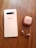 A smartphone placed on a wooden surface alongside a pair of wireless earbuds and their charging case. The phone is white with a triple camera setup. The earbuds and case are metallic copper, reflecting light.
