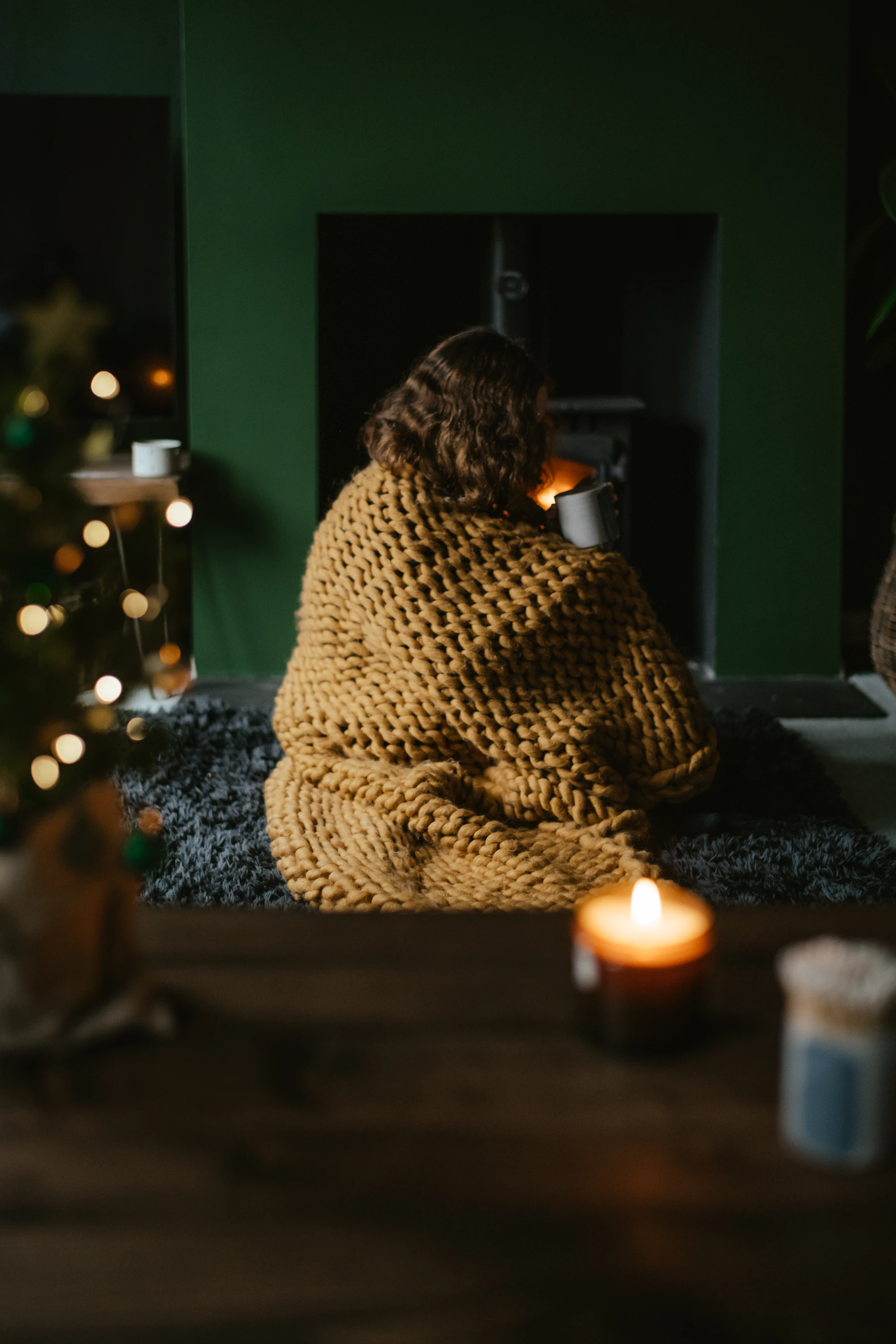 A person wrapped in a knit blanket in front of a fireplace