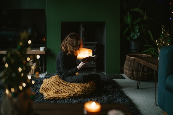 A cozy scene with a person sitting on a textured rug in front of a lit fireplace, reading a book. The setting includes a warm blanket, soft lighting, and a small decorated tree. The space is framed by dark green walls, a large woven basket, and a modern coffee table with a candle.