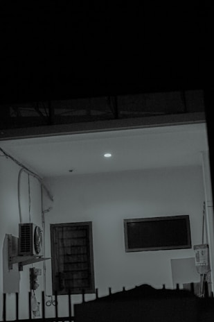 A dimly lit room with a visible air conditioning unit mounted on the wall. There is a closed wooden door next to a rectangular, unlit monitor or TV screen. The ceiling light casts a soft glow, and there are various wires and cables along the walls. A fence can be seen in the foreground.