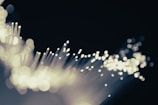 A close-up of fiber optic cables with glowing tips, creating a bokeh effect against a dark background. The scene appears abstract and dynamic as light seems to pulse through fine strands.