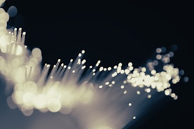 A close-up of fiber optic cables with glowing tips, creating a bokeh effect against a dark background. The scene appears abstract and dynamic as light seems to pulse through fine strands.