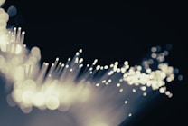 A close-up of fiber optic cables with glowing tips, creating a bokeh effect against a dark background. The scene appears abstract and dynamic as light seems to pulse through fine strands.