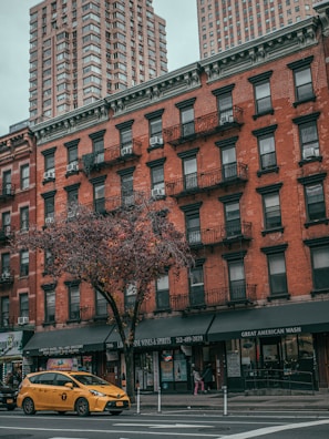 A bustling urban street scene featuring a red brick building with multiple floors and black metal fire escapes. The ground floor hosts various shops including a deli and liquor store. A yellow taxi is driving past along a tree-lined avenue, and high-rise buildings tower in the background under a cloudy sky.