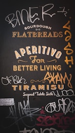 A wall mural featuring a mix of promotional text and graffiti. The text includes words like 'Sourdough Flatbreads,' 'Aperitivo for Better Living,' and 'Tiramisu Served Table Side.' Surrounding the promotional phrases are various graffiti tags in different styles. There is also a small sticker that reads 'Stop!! Uyghur Genocide.'