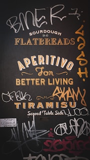 A wall mural featuring a mix of promotional text and graffiti. The text includes words like 'Sourdough Flatbreads,' 'Aperitivo for Better Living,' and 'Tiramisu Served Table Side.' Surrounding the promotional phrases are various graffiti tags in different styles. There is also a small sticker that reads 'Stop!! Uyghur Genocide.'