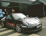 A sleek black sports car is parked on a paved area, featuring decals and sponsorship logos on its body. Three people stand nearby, with two men engaged in conversation and another person wearing a blue and white patterned hoodie. A large black tent is set up in the background, and there is a modern building with large glass windows behind them.