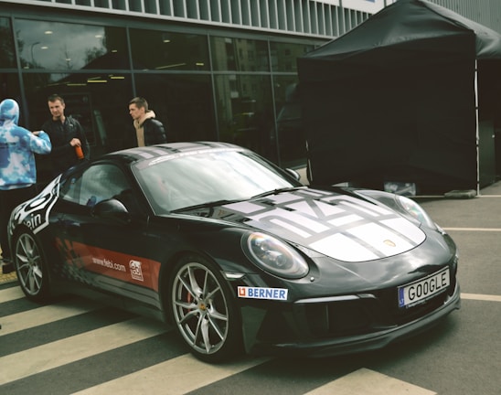 A sleek black sports car is parked on a paved area, featuring decals and sponsorship logos on its body. Three people stand nearby, with two men engaged in conversation and another person wearing a blue and white patterned hoodie. A large black tent is set up in the background, and there is a modern building with large glass windows behind them.