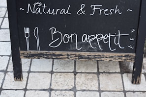 A black chalkboard sign with handwritten text displaying the words 'Natural & Fresh' and 'Bon appetit' alongside simple drawings of a fork and knife. The sign is set on a tiled stone floor.