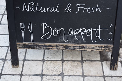 A black chalkboard sign with handwritten text displaying the words 'Natural & Fresh' and 'Bon appetit' alongside simple drawings of a fork and knife. The sign is set on a tiled stone floor.