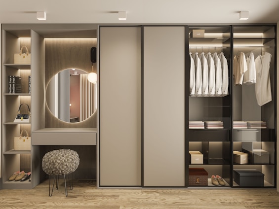 A modern and organized wardrobe with sliding doors, featuring a section with neatly hung shirts and a second section with folded clothes and boxes. To the left, there is a built-in vanity area with a circular mirror, small vanity table, and an elegant stool with woven fabric. The shelves contain handbags, shoes, and decor items. Soft lighting illuminates the space, highlighting the minimalist design.