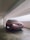 A red car parked in a covered parking garage. The car's headlights are on, and the license plate is visible. The concrete garage has a clean, minimalistic design with white walls and exposed pipes on the ceiling.