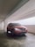 A red car parked in a covered parking garage. The car's headlights are on, and the license plate is visible. The concrete garage has a clean, minimalistic design with white walls and exposed pipes on the ceiling.