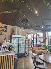 A cozy cafe interior featuring a wooden floor, with decorative greenery and lights adorning the ceiling. A refrigerated display case showcases floral arrangements. To the side, there's a coffee counter with various syrups and a seating area with a tan couch and round tables. Large windows allow natural light to enter, and a shelf displays snacks and drinks.