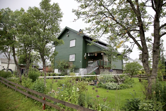 A green two-story wooden house is surrounded by lush greenery and trees. The garden is well-maintained with a variety of plants and flowers, and a wooden fence runs along the front. The sky is overcast, adding a serene and peaceful ambiance.