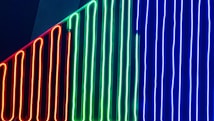 Vertical neon lights in red, green, and blue are arranged in parallel lines against a dark background, creating a vibrant and modern aesthetic.