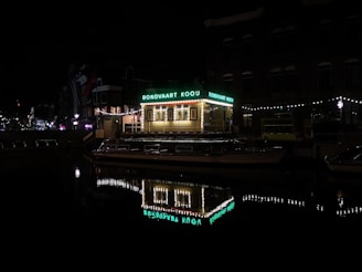 A nighttime scene featuring a brightly lit canal boat with a roof sign reading 'RONDVAART KOOIJ'. The boat is docked along a calm waterway reflecting the lights from the boat and surrounding buildings. String lights adorn the nearby structure and create a festive atmosphere. The surrounding buildings are dimly lit, contributing to the quiet and serene night ambiance.