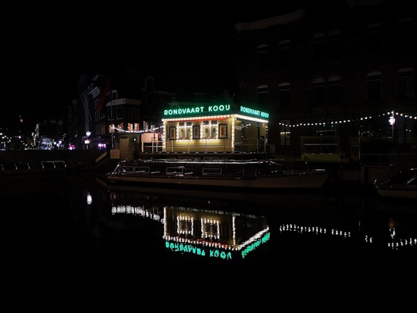 A nighttime scene featuring a brightly lit canal boat with a roof sign reading 'RONDVAART KOOIJ'. The boat is docked along a calm waterway reflecting the lights from the boat and surrounding buildings. String lights adorn the nearby structure and create a festive atmosphere. The surrounding buildings are dimly lit, contributing to the quiet and serene night ambiance.