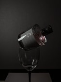 A dark, moody setting features a perfume bottle delicately balanced on the rim of a wine glass. The bottle is adorned with droplets of water, reflecting minimal light, creating a dramatic and elegant vibe. The background is a deep grey, adding to the sophisticated ambiance.