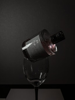A dark, moody setting features a perfume bottle delicately balanced on the rim of a wine glass. The bottle is adorned with droplets of water, reflecting minimal light, creating a dramatic and elegant vibe. The background is a deep grey, adding to the sophisticated ambiance.