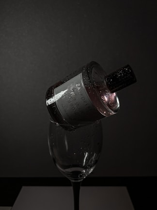 A dark, moody setting features a perfume bottle delicately balanced on the rim of a wine glass. The bottle is adorned with droplets of water, reflecting minimal light, creating a dramatic and elegant vibe. The background is a deep grey, adding to the sophisticated ambiance.